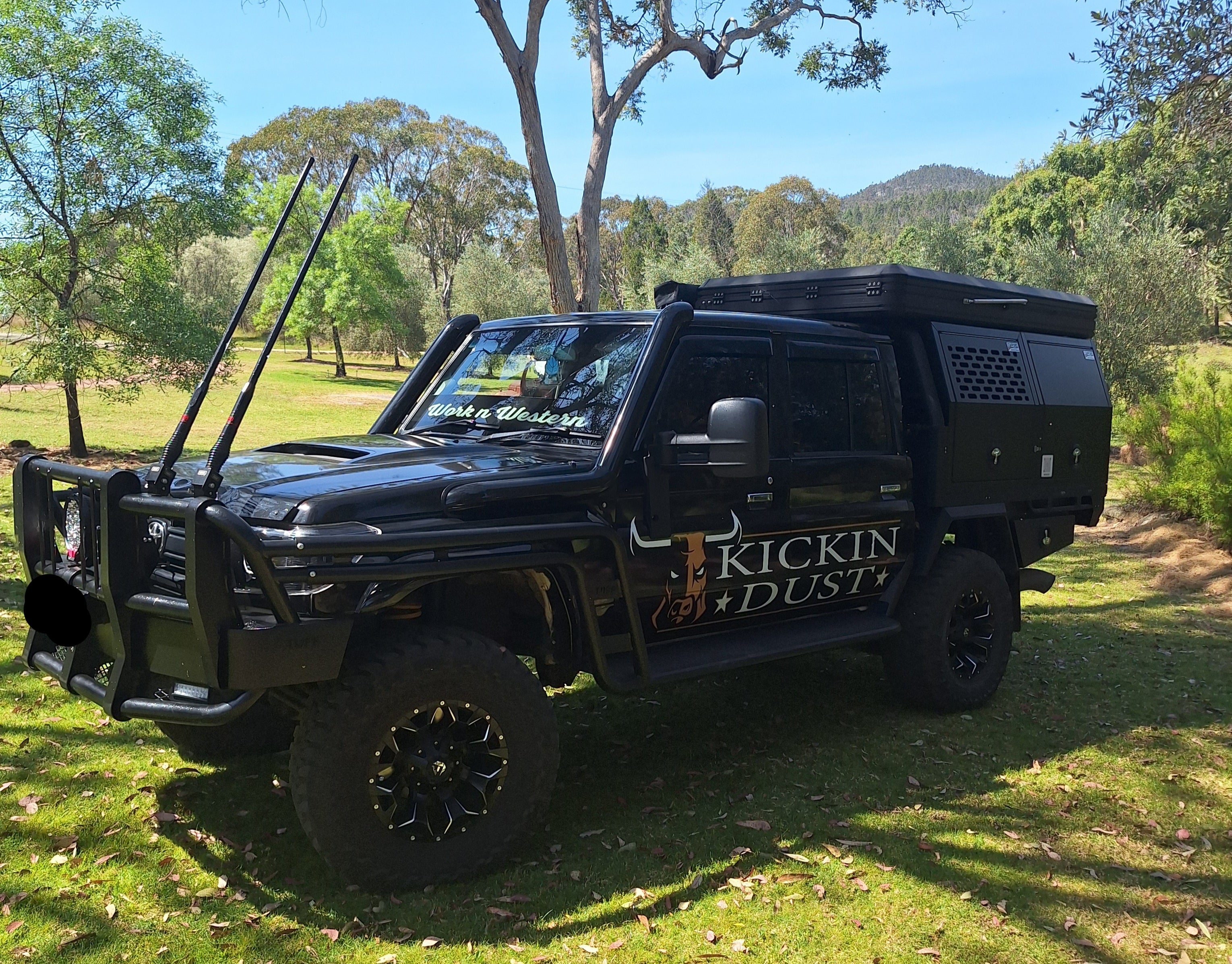Black off-road vehicle with 'Kickin Dust' branding parked on grass with trees in the background
